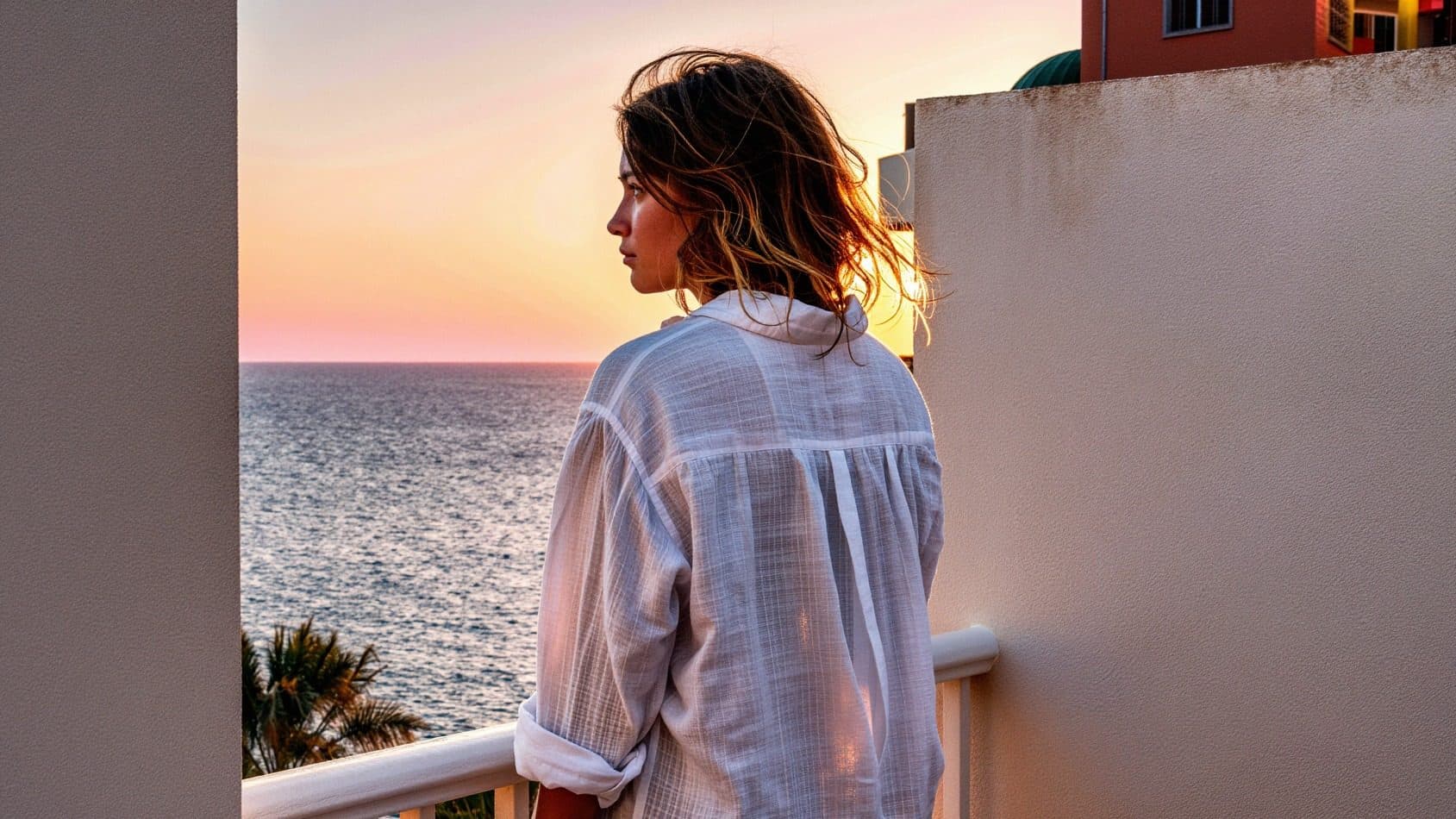 A woman seen from behind on her hotel balcony in Ibiza, looking out at the sea at sunset