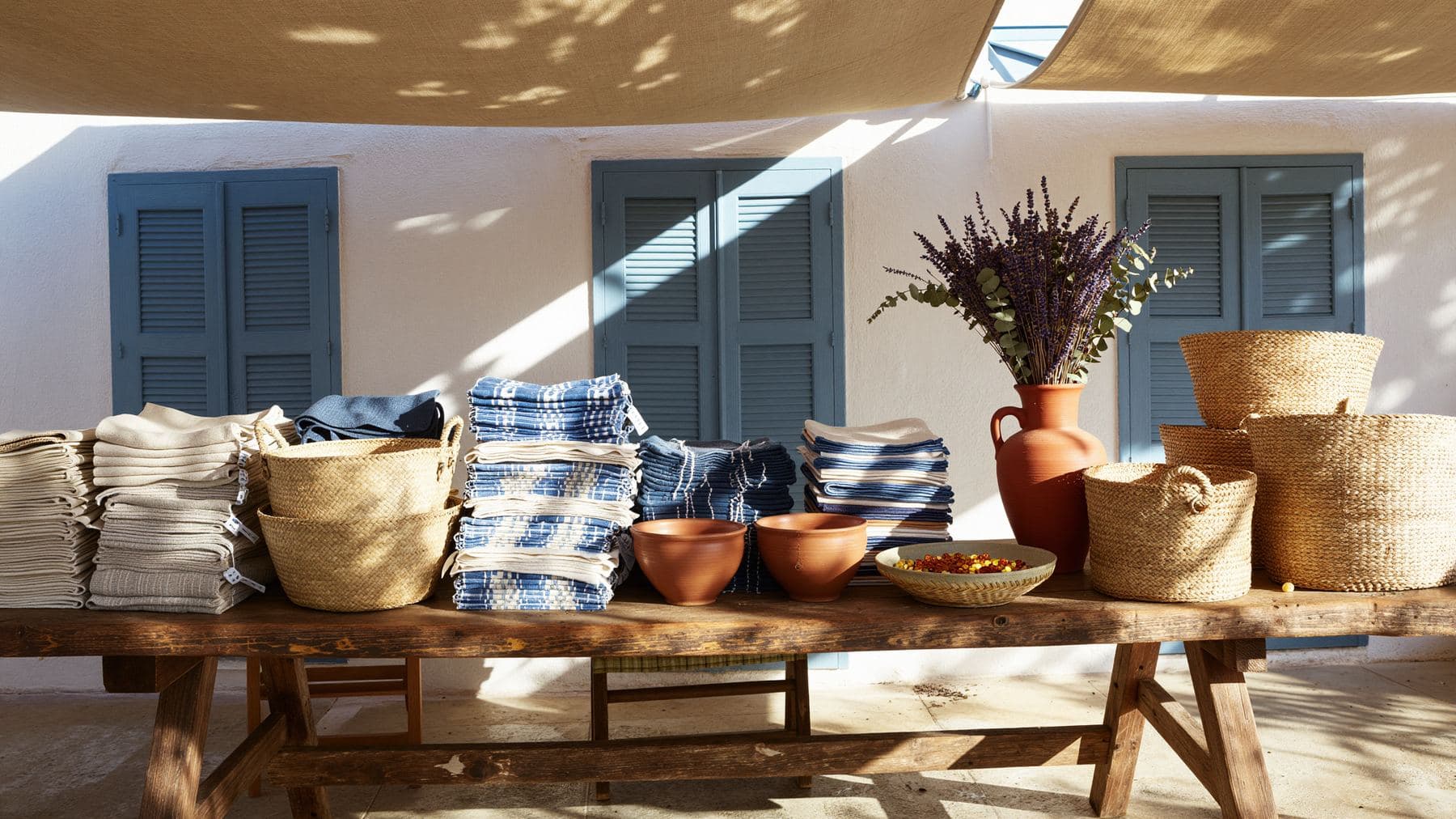 A rustic wooden market table in dappled shade with stacked linen, straw baskets and ceramic bowls