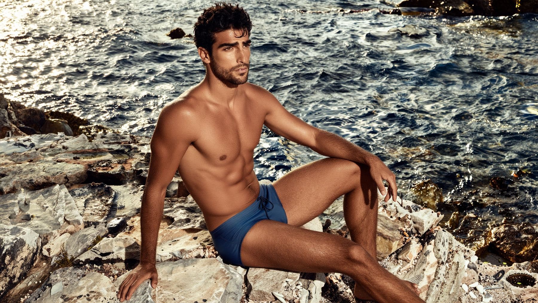 A handsome Mediterranean man, shirtless and wet from a swim, sitting on the rocks at the edge of the sea in an Ibiza cove at golden hour