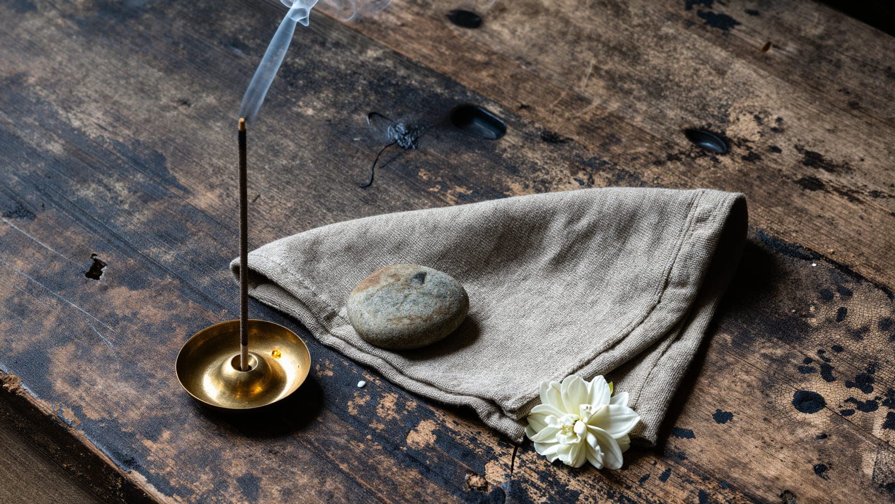A lit incense stick in a brass holder on dark weathered wood, beside folded linen, a smooth stone and a white jasmine flower