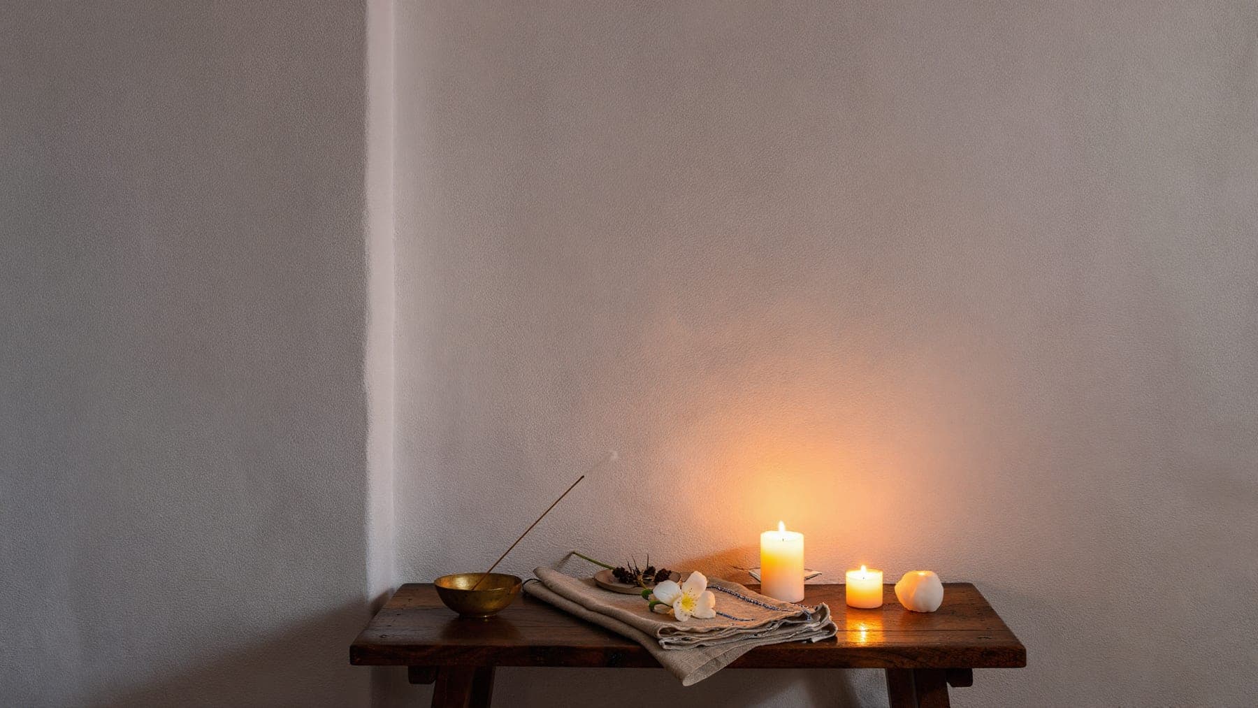 A small altar with a lit incense stick, a brass bowl and three candles in a whitewashed room at dusk